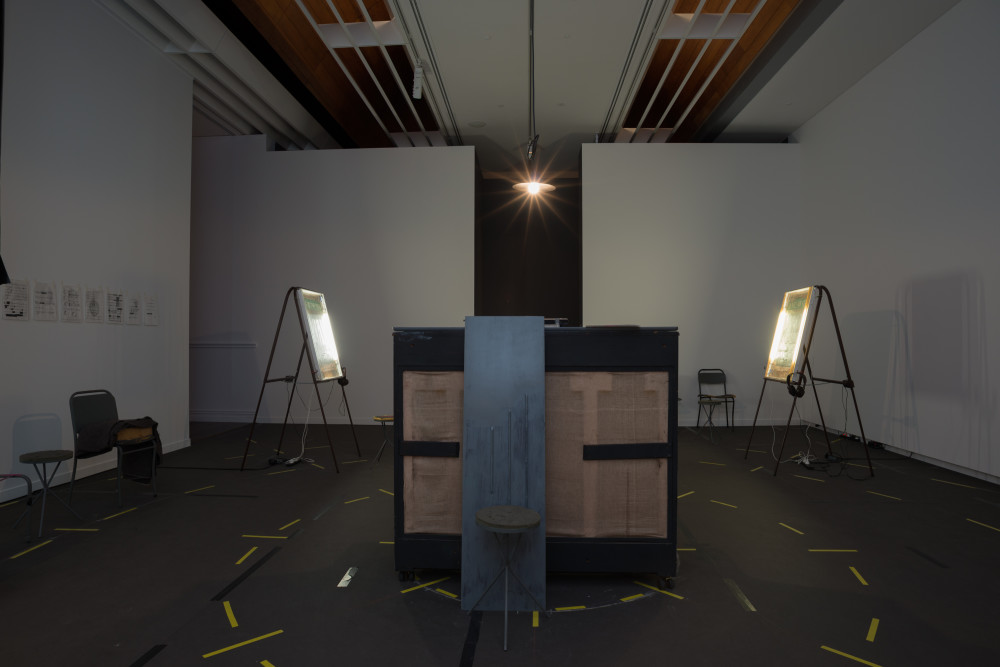 The image shows a large dimly lit gallery space. In the centre of the space stands an upright piano, facing away from the viewer so that its unpainted wooden back is exposed. At the other end of the room on each side of the piano stand two easels, which each hold neon lightboxes with headphones next to each. The floor of the space is covered with pieces of yellow tape which circle the installation multiple times. Some small drawings in plastic protectors are pinned to the left hand gallery wall, with chairs below them. 