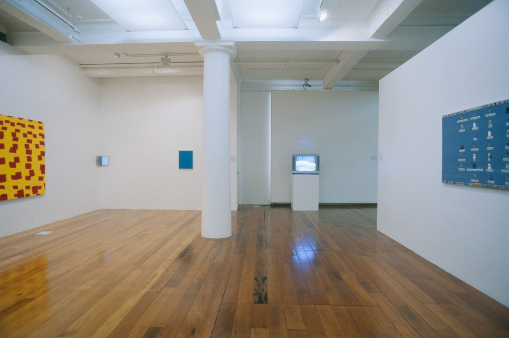 The image shows a white gallery space with a polished wooden floor. On the left is a painting made up of dark red and bright yellow squares. Next to it are two smaller blue artworks, not clearly visible from the photograph's distance. In the middle of the room is a tall white column, and to the right of this is an analogue television on a plinth playing a video. On the far right a blue painting hangs on the wall. 
