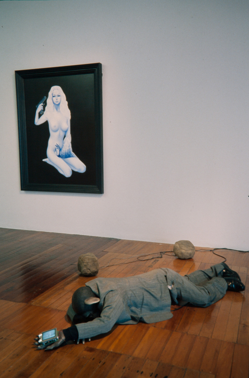 On the back white gallery wall hangs a realistic painting of a unnaturally pale, blue-tinged nude woman sitting down in a black void space. She stares at the viewer while holding a gun to her head. In the foreground, a lifelike replica of a chimp is sprawled across a polished wooden floor, dressed in a dark grey business suit. He is positioned face down and holds a device with a screen on it in one outstretched hand. 