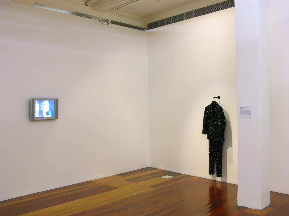 On the left hand white gallery wall, a small rectangular flatscreen television plays a video depicting a person blowing up a balloon one handed. On the right wall hangs a dark black suit covered in little pointed studs.