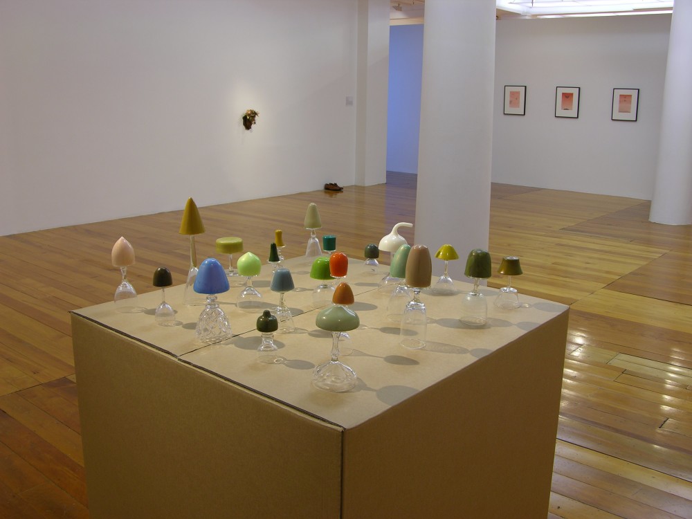In the foreground is a large square cardboard plinth with multiple tiny glass mushroom sculptures in various colours arranged upon it. In the background, two-dimensional framed artworks are hung on white gallery walls with a column in the middle of the room. 