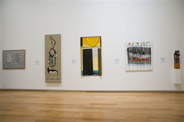 The image depicts a wall in one of the ground floor galleries at Auckland Art Gallery. On the far left hangs an artwork filled with horizontal koru patterns in alternating black and white. Next to this hangs a long narrow panel with a brown background, depicting a stylised figure in black. In the centre hangs a loose canvas with an orange and yellow section at the top depicting a sun, and an ocean view broken up by sections of solid black. On the left hangs a white artwork with black and white vertical lines all over it, with two horizontal blue lines on either side.