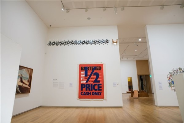 One of the ground floor galleries at Auckland Art Gallery is depicted. On the left wall hangs a red and blue work which reads '1/2 price cash only' on loose canvas. Above it, multiple identical signs bearing the letter 'O' are hung together on an angle. A tall doorway to the right of these works looks into the adjoining galleries. 
