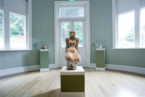 The image depicts a rounded bay room in Auckland Art Gallery. In the centre of the room a carved sculpture of a curvy, stylised person half crouches on a plinth. Behind the sculpture, two pieces of pottery sit in glass vitrines. Multiple windows surround these artworks, looking out onto Albert Park.