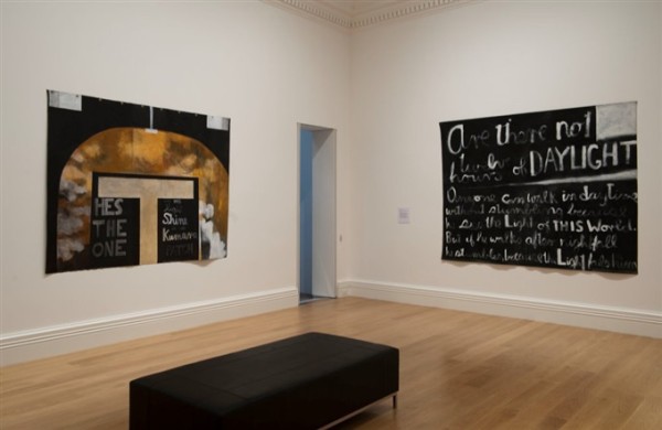 The image depicts a corner of a room in Auckland Art Gallery. On the left hangs a loose canvas with a black background, with a T shape in the middle in beige and an arc of golden paint surrounding it. 'He's the one' and 'may his light shine' are written in white paint underneath the T shaped cross. On the right wall hangs another loose canvas with a black background, which is filled with lettering written with white paint. 