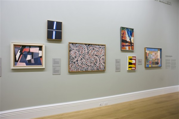 The image depicts a wall in Auckland Art Gallery, with multiple framed artworks hung at different heights next to each other along the wall. In the centre is a work filled with swirling kowhaiwhai inspired patterns in black, white and red. The other works are a mixture of abstract geometric shapes and figurative but geometric landscapes. 