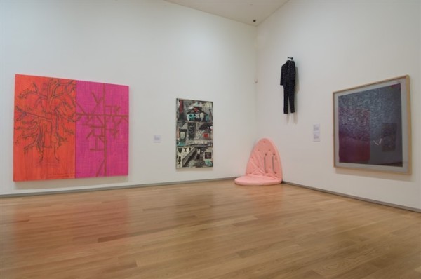 One of the ground floor galleries at Auckland Art Gallery is depicted. On the right hand wall hangs a large pink and orange canvas with a spindly tree painted onto it. To its right hangs a narrow figurative painting depicting suburban scenes. In the corner of the room, a salmon pink circular mattress is half propped up against the wall. On the right hand wall, above the mattress hangs a man's suit with sharp studs all over it. On the far right hangs a large framed painting depicting a tree.
