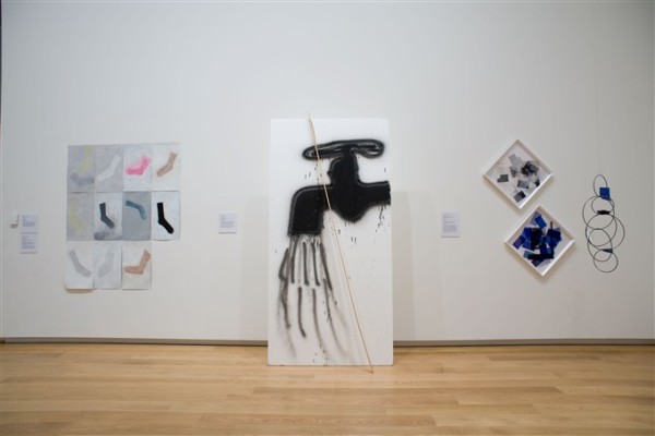 The image shows a wall in the ground floor galleries at Auckland Art Gallery. On the left is a series of photographs of socks, arranged in a grid. In the centre a tall board is propped against the wall, with a large black tap spray painted onto it and emitting black lines of 'water'. On the right hang two diamond shaped works filled with a random assortment of shapes.