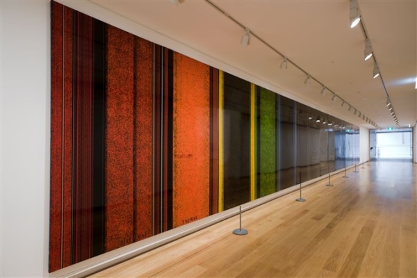 The image is taken from an angle showing the length of a corridor with a very long artwork stretching down the whole wall. The work is a lacqured, glossy panel with a series of fine lines running vertically down it, with sections of bright colours of the rainbow dotted in between.